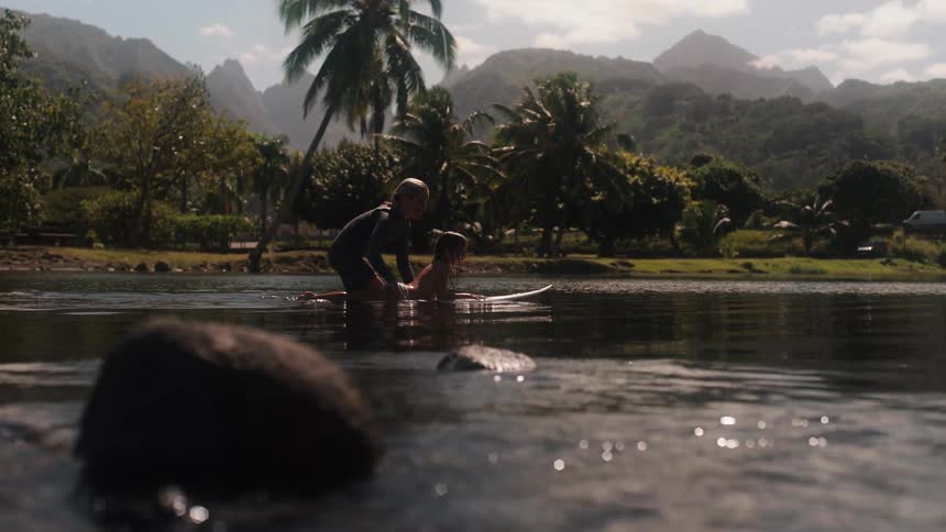 A woman teaches a young child to surf in the calm, tropical waters of a lush, mountainous island
