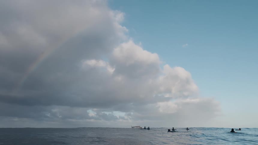 A slow-motion reveal from the ocean's surface shows a group of surfers waiting for waves under a magnificent rainbow