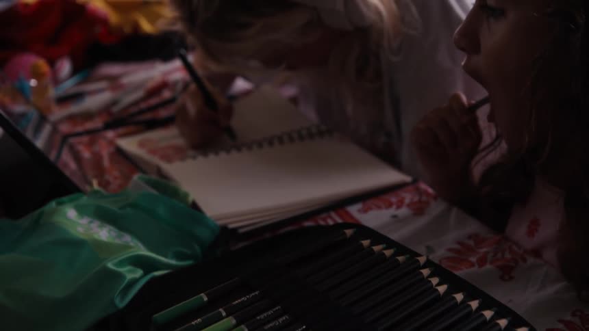 A close-up reveals a large pencil case before tilting up to show two young sisters drawing together in a sketchbook