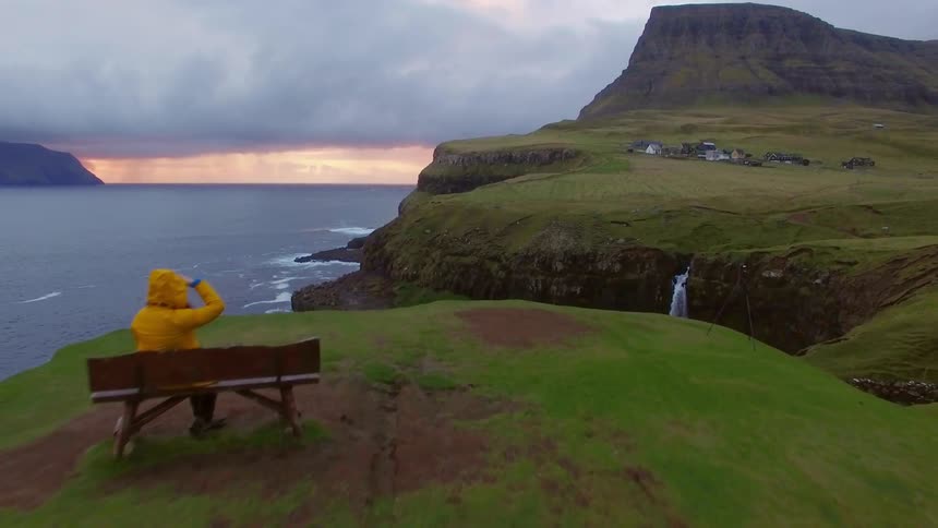 A woman sits down on a bench looking out toward a village and the ocean