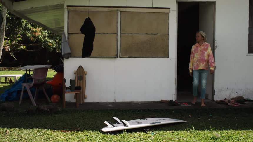 A barefoot girl in a tie-dye hoodie slowly walks out of a surf shack with her surfboard on the lawn