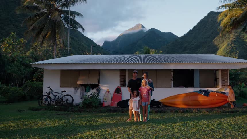 A slow zoom out reveals a beautiful family portrait in front of their tropical island home with lush green mountains in the background