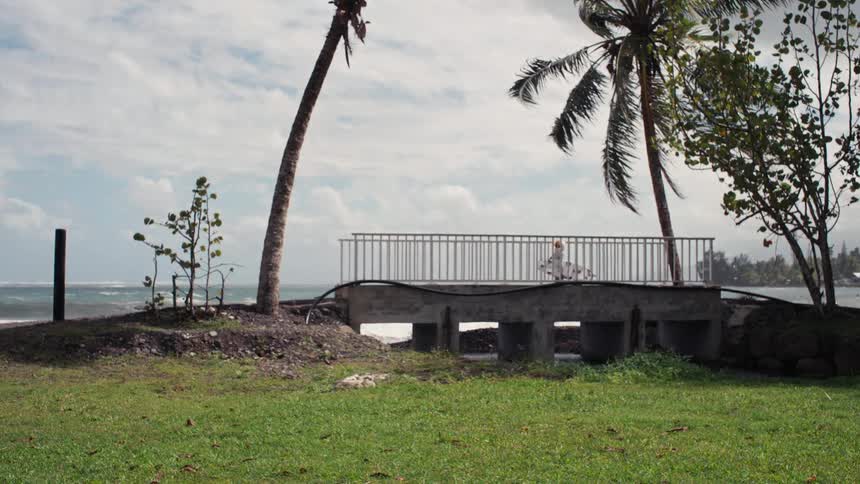 A young surfer carries their surfboard while walking across a small bridge next to the ocean on a tropical island