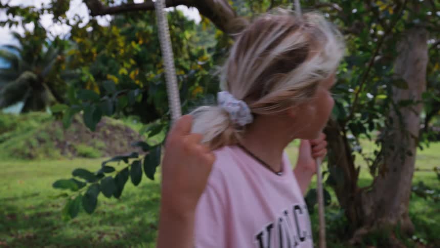 A contemplative young blonde girl gently sways on a rope swing in a lush, green tropical yard in slow motion
