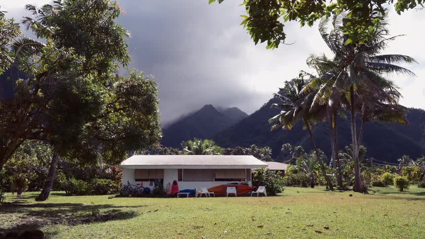 A peaceful tropical home with kayaks and surfboards rests in a lush yard as storm clouds brew over distant mountains