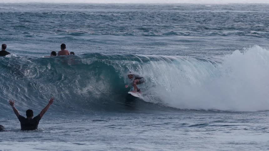 A skilled surfer expertly rides inside the barrel of a powerful ocean wave in super slow motion as other people watch from the water