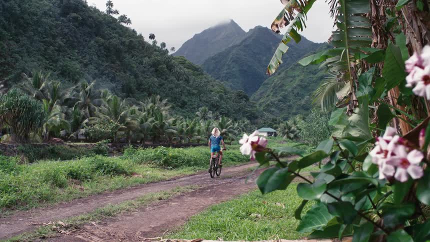 A young blonde girl rides her bicycle down a dirt path through a lush tropical valley surrounded by green mountains and palm trees