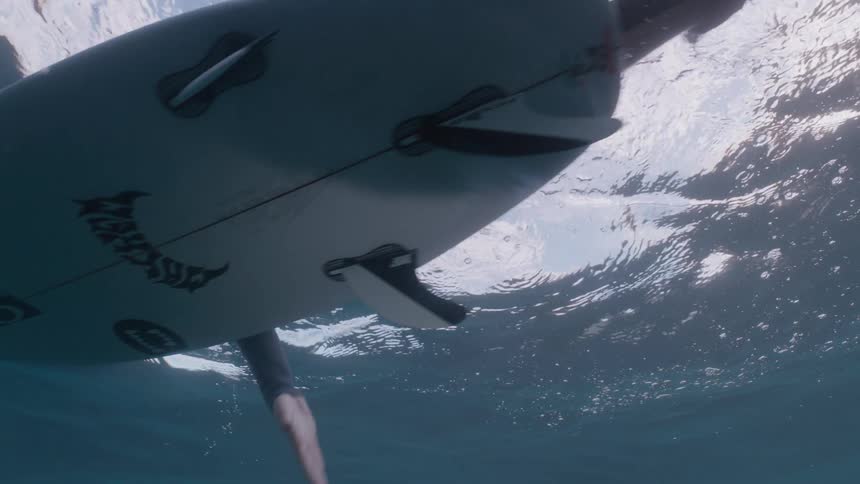 A serene underwater view captures a surfer in slow motion paddling and waiting for a wave from below the ocean's surface