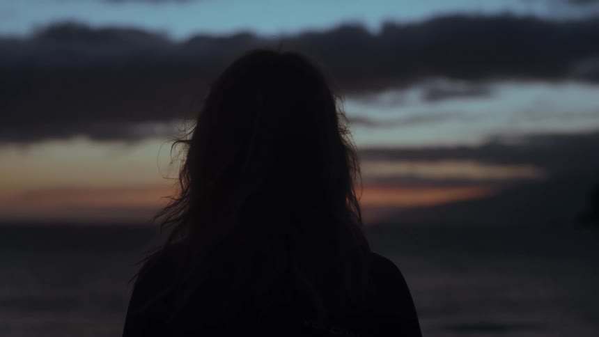 A static shot shows the silhouette of a woman from behind as she peacefully watches the sunset over the ocean
