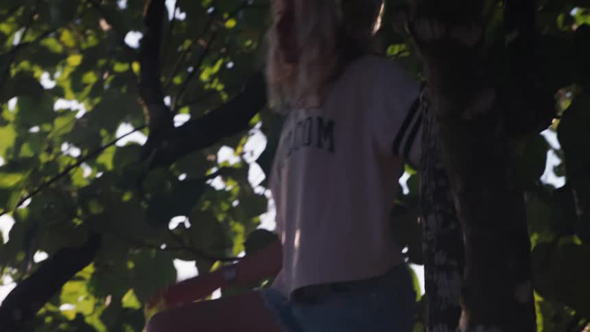 A young blonde woman carefully climbs down a leafy tree in slow motion, backlit by the warm golden hour sun