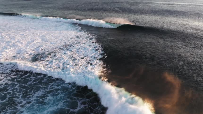 An aerial slow-motion view captures a lone surfer expertly riding inside the barrel of a perfect ocean wave as the golden sunset reflects off the sea spray