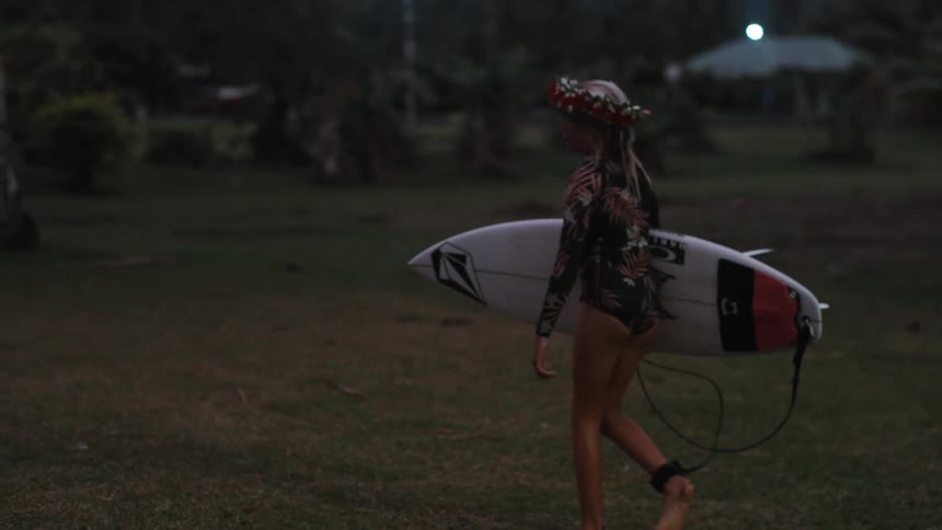 A young surfer girl with a flower crown carries her surfboard across a grassy field at dusk in slow motion