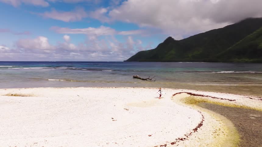 An aerial orbit shot of a woman standing on a remote white coral sandbar surrounded by a tropical ocean and lush green mountains