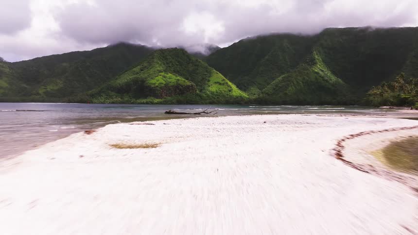 An exhilarating aerial drone flight skims over a shallow coral reef towards a lone surfer on a sandbar, set against a backdrop of majestic green mountains