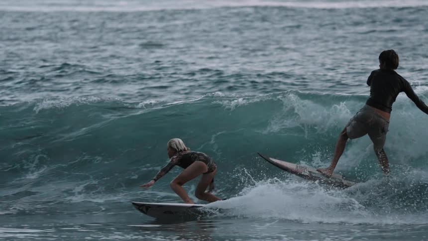 A young girl and boy skillfully surf a rolling ocean wave together in stunning slow motion while an adult looks on