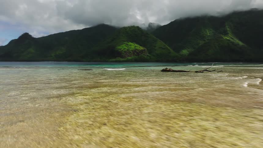 An aerial shot follows a woman with a surfboard walking along a pristine white sandbar towards a lush, mountainous tropical island