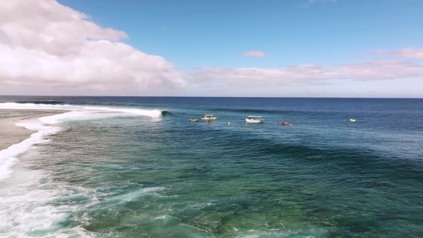 Boats and surfers await the next set of perfect, turquoise waves breaking over a shallow reef on a beautiful day