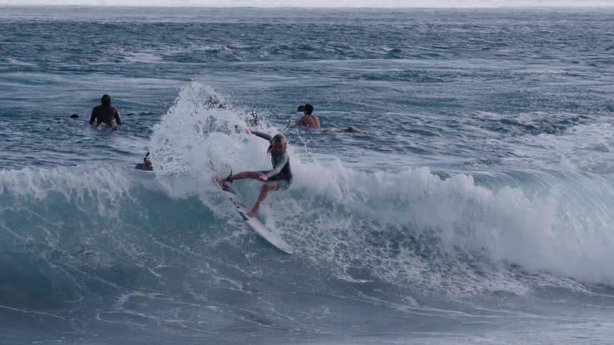 A surfer skillfully carves a large ocean wave in super slow motion as other swimmers watch from the background