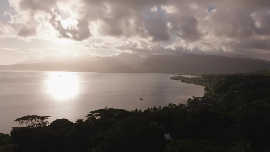 A majestic aerial view captures a serene tropical bay at sunset, where the sun breaks through dramatic clouds to reflect on the calm water