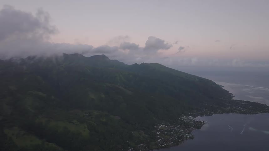 A majestic aerial view of a lush green tropical island with cloud-covered mountains and a coastal town at dusk