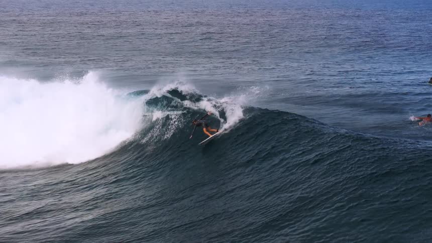 A surfer skillfully rides a massive ocean wave in breathtaking slow motion as other surfers watch
