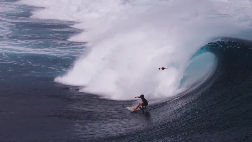 A surfer skillfully rides inside the barrel of a massive ocean wave, closely followed and filmed by a drone in stunning slow motion