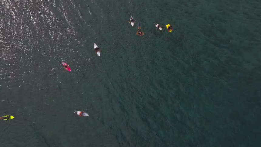 An aerial top-down view shows a group of surfers patiently waiting for waves in the calm ocean water