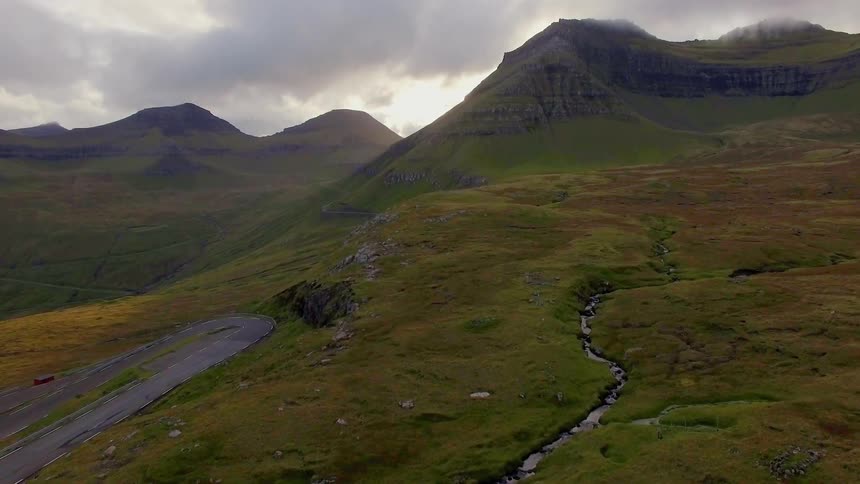 A creek runs down a hillside near a winding road on a cloudy day