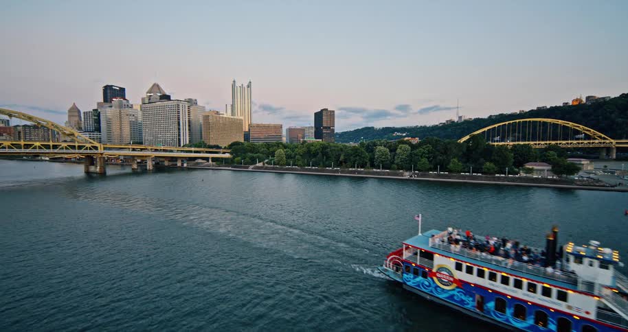 An aerial view of a sightseeing riverboat cruising along the river past the Pittsburgh city skyline at twilight