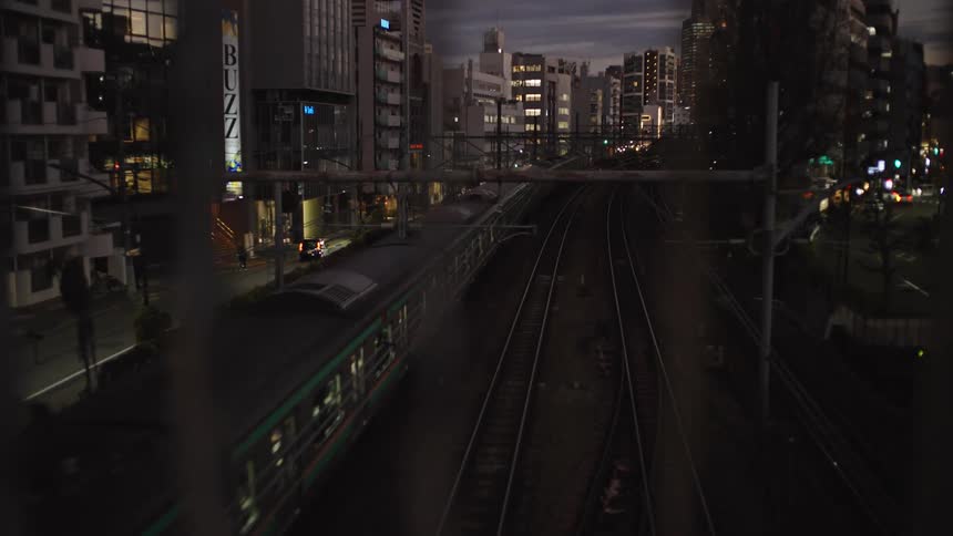 Through a fence, a commuter train travels through the city of Tokyo at dusk