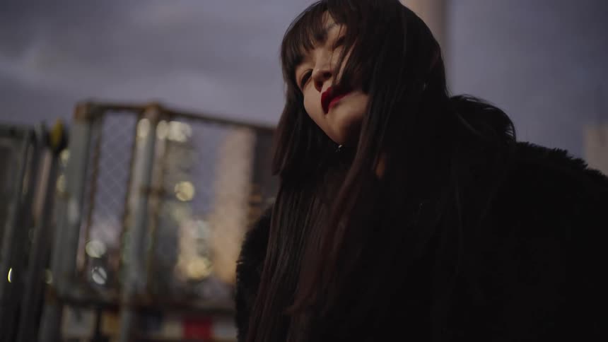 A moody close-up of a stylish Japanese woman with red lipstick as the city lights come on behind her at dusk