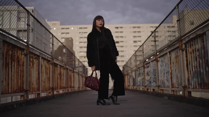 A fashionable Asian woman in a black outfit and fur coat poses confidently on a rusty urban overpass at dusk