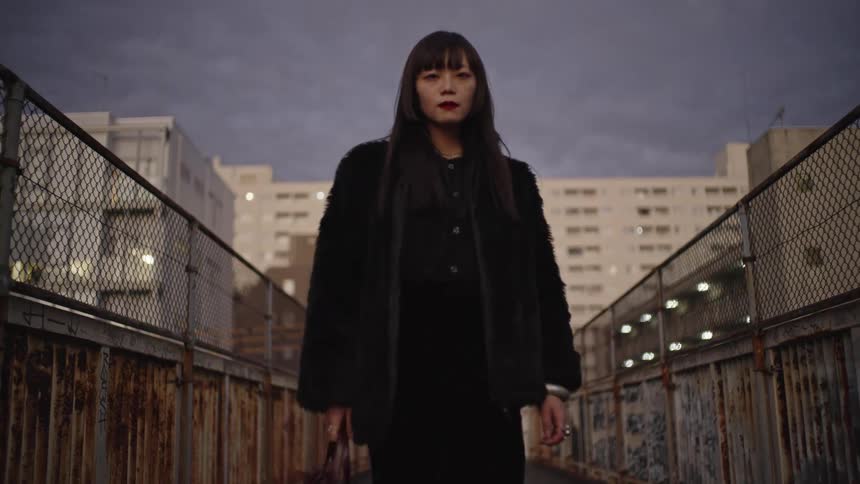 A stylish Japanese woman confidently walks down a gritty urban overpass towards the camera in slow motion at dusk