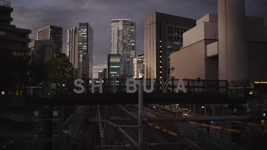 A static shot of the Shibuya sign over train tracks with the Tokyo cityscape in the background at dusk