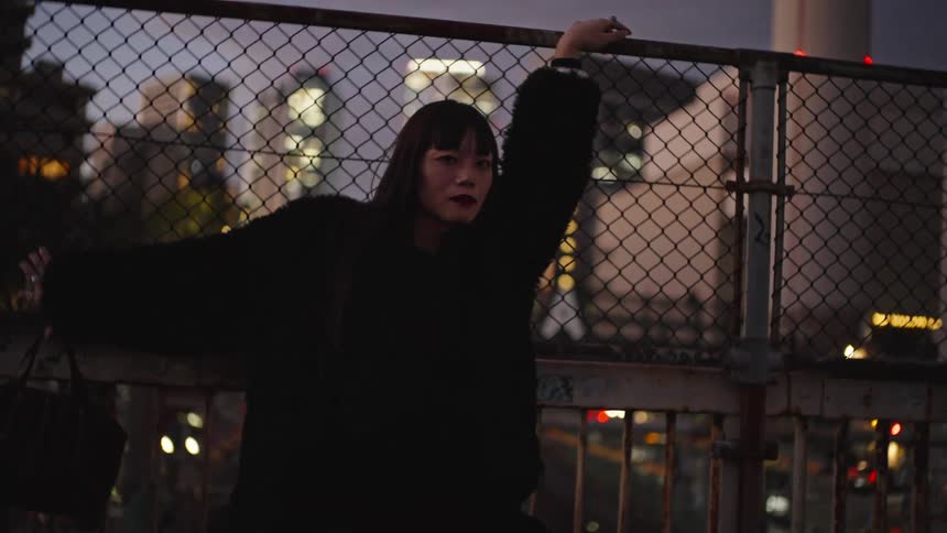 A stylish young Asian woman poses against a chain link fence on an urban overpass at night with the city lights in the background