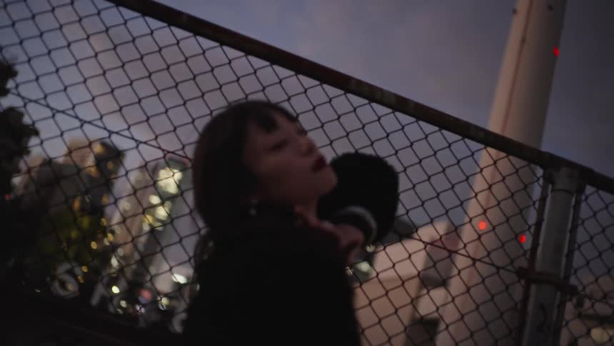 A stylish Asian woman poses melancholically against a chain-link fence, with the city lights of Tokyo blurred in the background at dusk