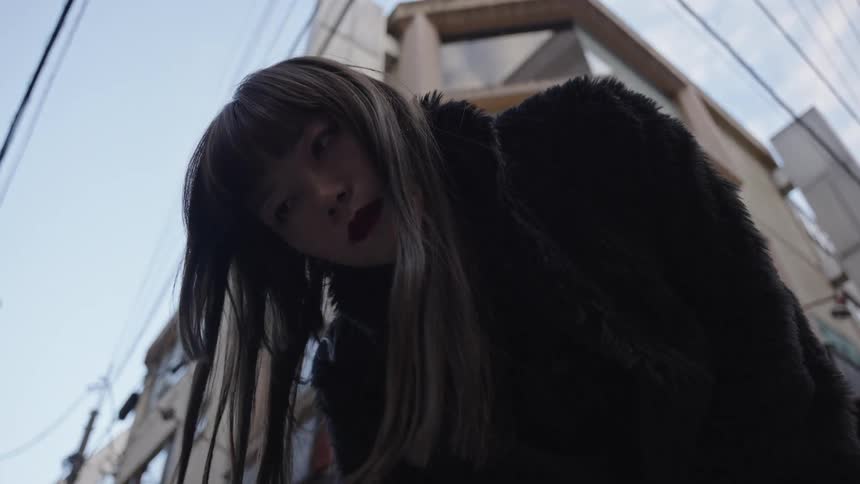 A stylish Japanese woman in a black fur coat looks down menacingly at the camera from a low angle on a city street