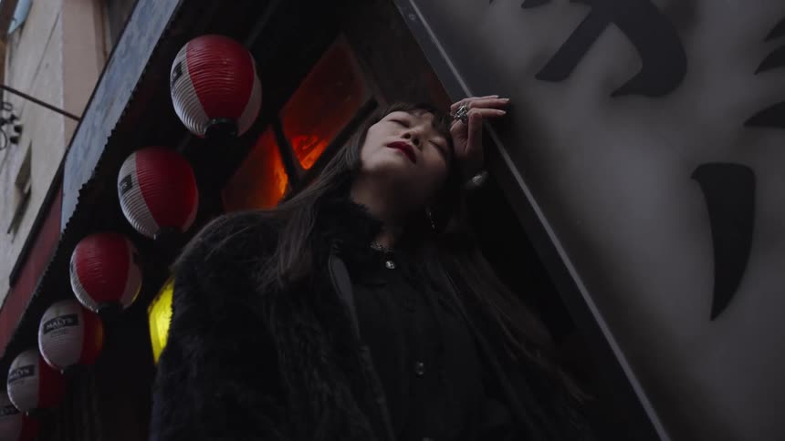 A fashionable Asian woman in a black fur coat leans against a city sign with her eyes closed, framed by traditional lanterns