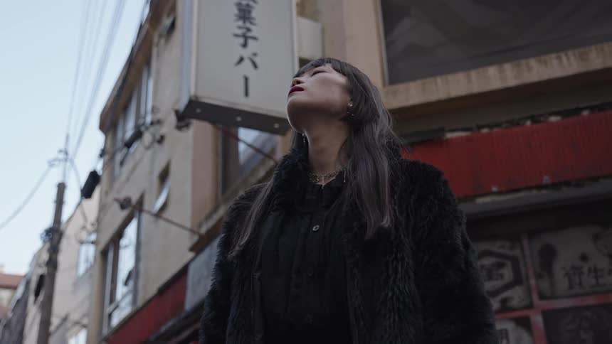 A thoughtful young Japanese woman in a black fur coat stands pensively in an urban alley, looking up towards the sky