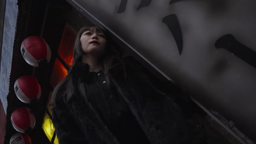 A woman in a black fur coat stands under a Japanese sign and lanterns in a moody, low-angle shot
