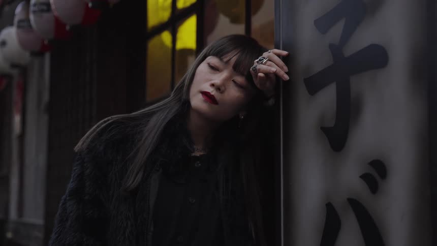 A contemplative and stylish Asian woman with red lipstick leans against a sign on a moody city street in Japan