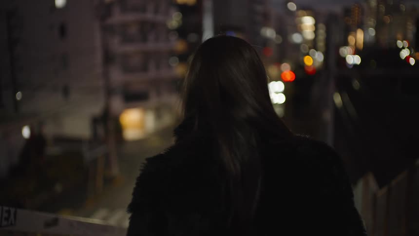 A stylish woman looks back over her shoulder with a pensive expression on a city overpass at night, then turns away as the scene blurs