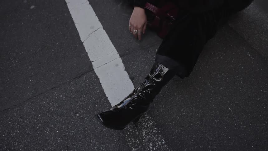 A high-angle detail shot of a stylish person's shiny black heeled boot as they sit on the asphalt of a city street