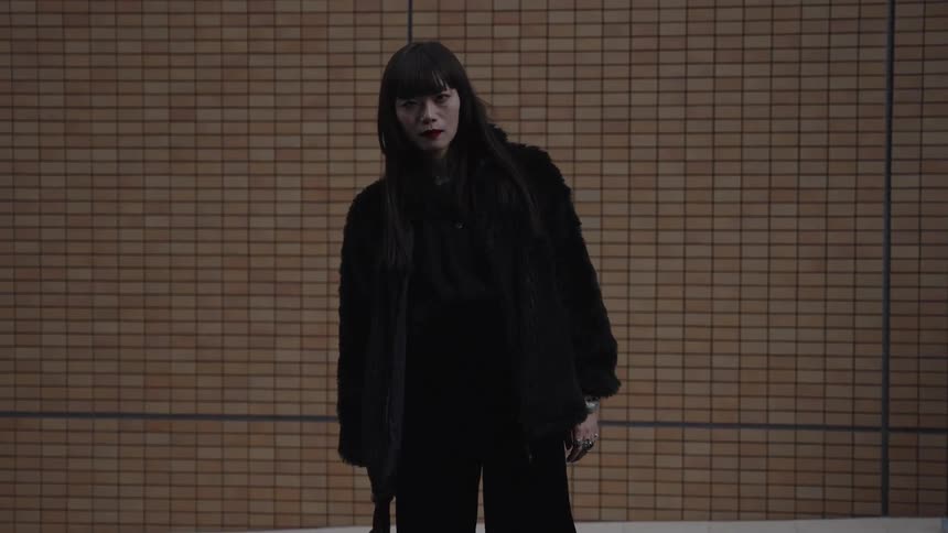 A stylish Japanese woman in a black fur coat and red lipstick stares intently at the camera against a tiled wall