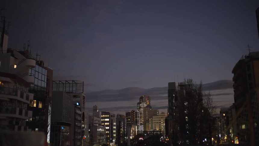 A quiet evening settles over a bustling city in Japan, with building and traffic lights glowing against the twilight sky