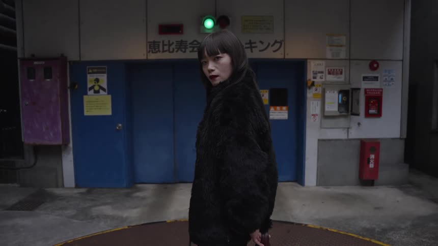 A fashionable Japanese woman in a black furry coat turns slowly and gives an intense look to the camera in a moody urban alleyway