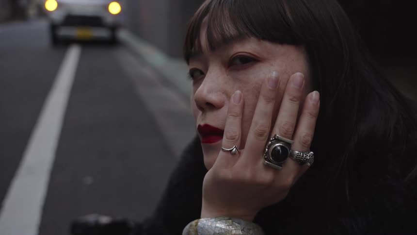 A close-up cinemagraph of a pensive woman with bold jewelry and red lipstick on a city street as a car drives by