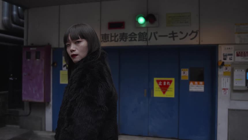 Slow motion portrait of a fashionable Japanese woman in a black fur coat staring at the camera in front of a parking garage entrance