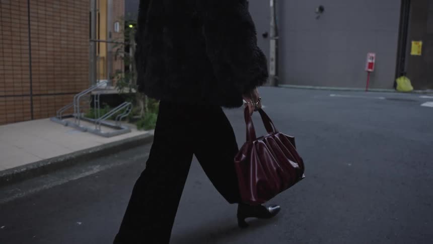 A stylish woman in a black faux fur coat and heeled boots walks down an urban city street in slow motion