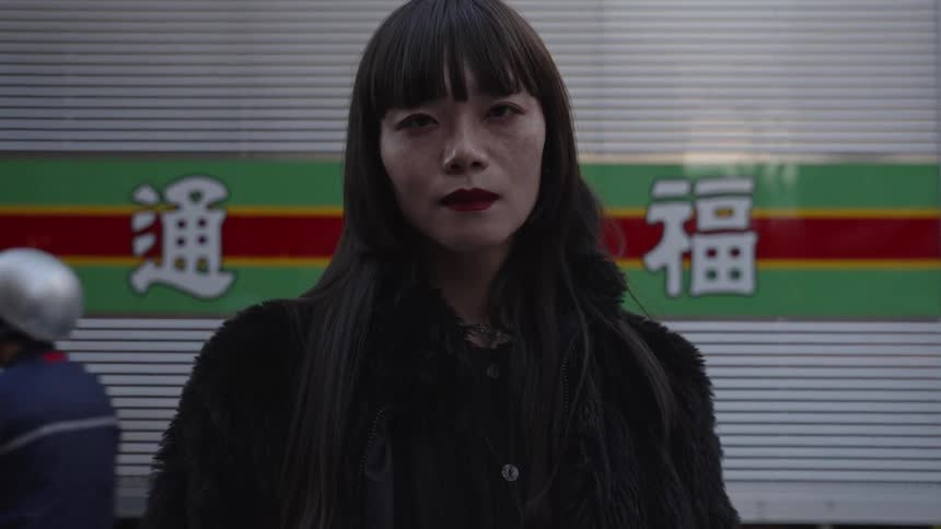 A slow-motion close-up portrait of a stylish Japanese woman with red lipstick staring intently at the camera on a city street